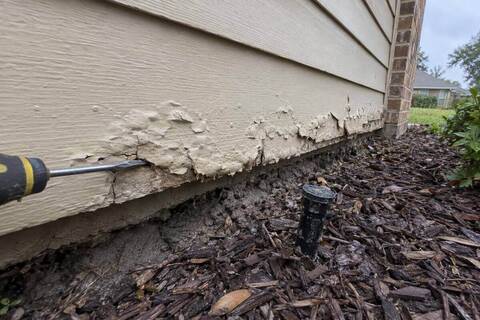 Swollen and flaking hardboard lap siding at the bottom of a wall above a flower bed