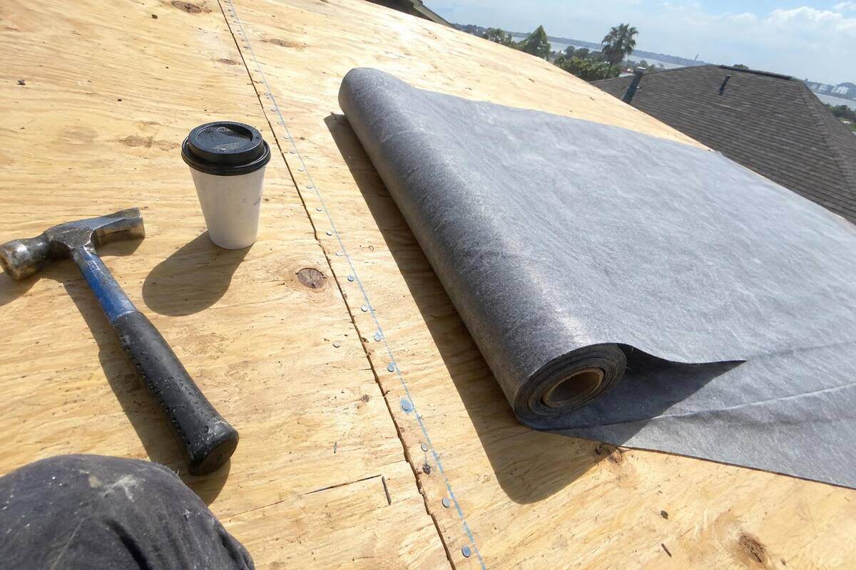 Close view of plywood roof decking with a tight row of nails along a chalk line on a rafter and a roll of synthetic underlayment beside it