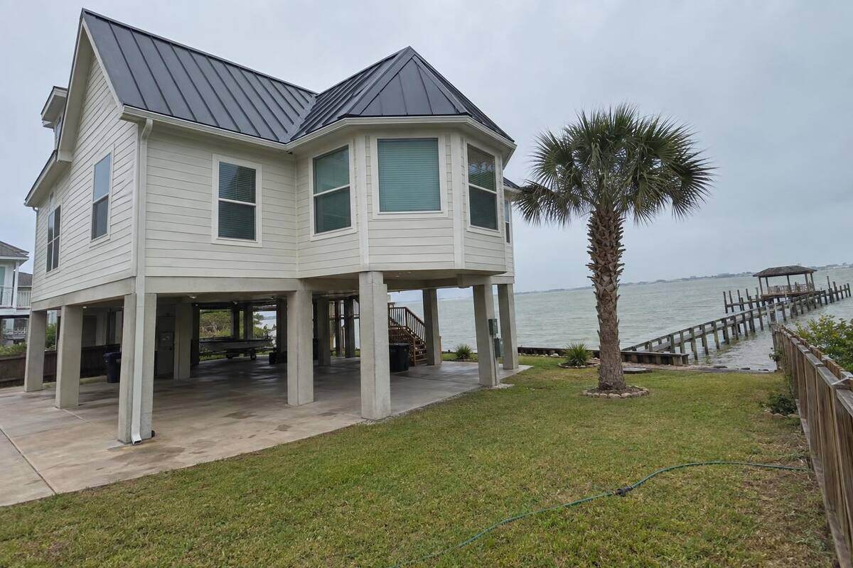 Charcoal standing-seam metal roof on a raised two-story house on pilings with Galveston Bay water visible beyond the yard